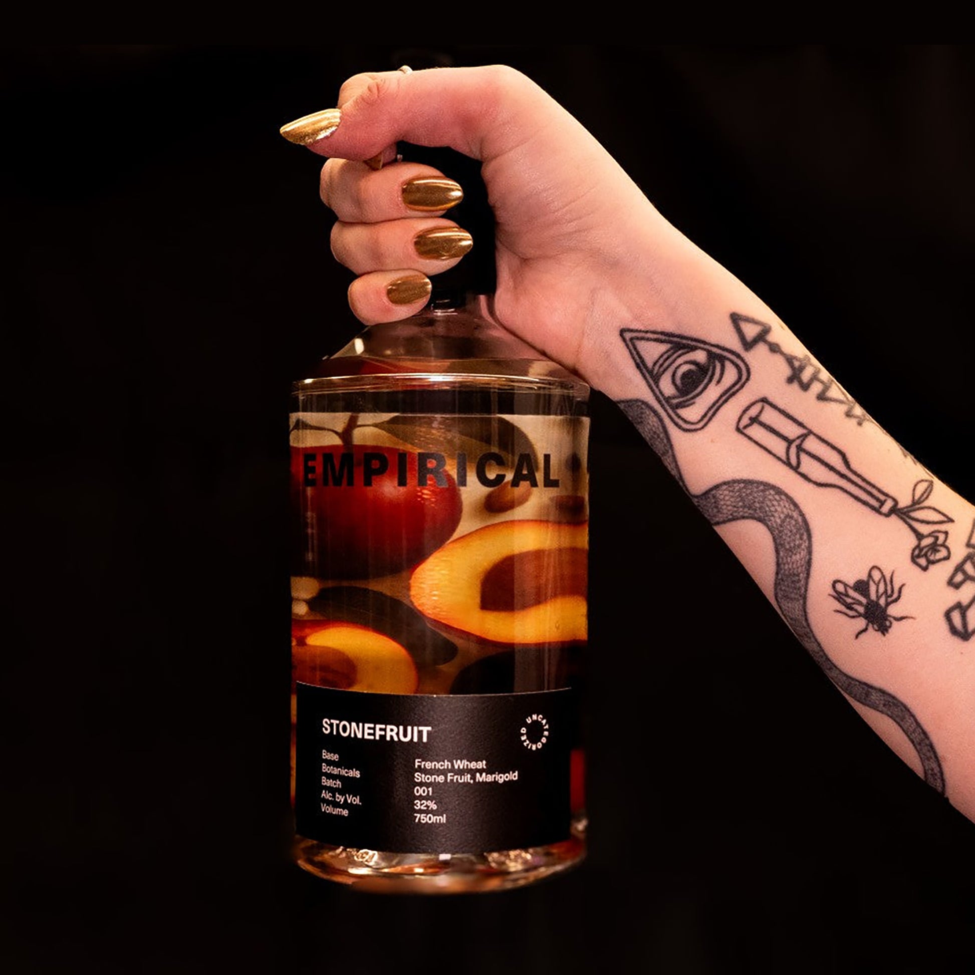 Hand with gold nails holding the Empirical Stonefruit bottle on a black background