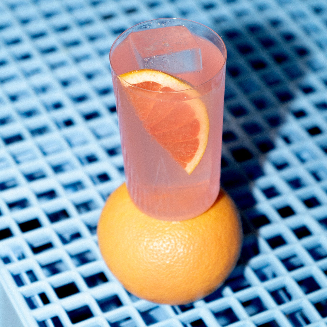 Empirical Ayuuk Paloma in a tall glass perched on a grapefruit, on a blue grid surface