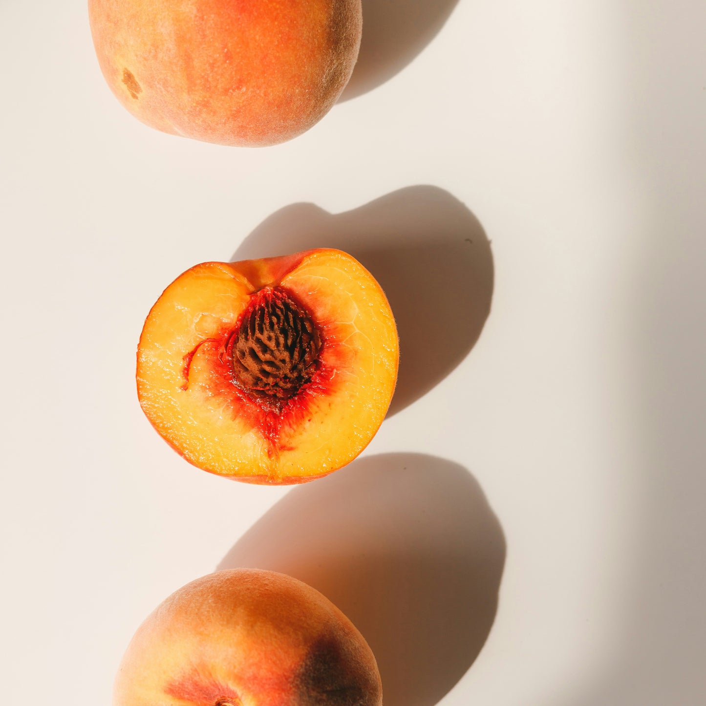 Three peaches arranged on a sunlit white surface, one halved to show the pit